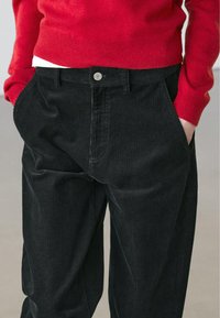 Pantalon en velours côtelé noir avec des rainures verticales, des poches avant, et une fermeture à bouton métallique, assorti à un pull rouge.