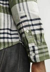 Camisa de franela a cuadros verdes y grises con accents en negro, que presenta un puño abotonado y una tela texturizada. Denim oscuro debajo.