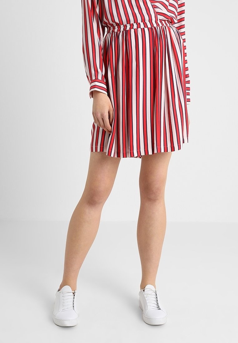 Personne portant une jupe et un haut à rayures verticales rouges, blanches et noires, associées à des baskets blanches, debout devant un fond uni.