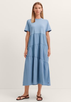 Frau steht und trägt ein hellblaues gestuftes Midikleid mit kurzen Ärmeln und braunen flachen Sandalen vor einem einfarbigen Hintergrund.