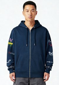 Sweat à capuche zippé marine avec une grande poche avant. Présente des motifs colorés brodés sur les manches, incluant des fleurs et des accents de texte.