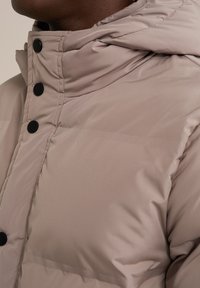 Béžová puffa bunda s kapucňou, polstrovanou textúrou a čiernymi patentkami pozdĺž prednej časti. Hladká tkanina s jemným leskom.