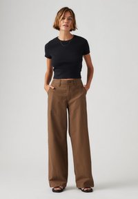 Schwarzes, kurzärmliges, figurbetontes T-Shirt kombiniert mit hochgeschnittenen, weiten braunen Hosen. Das Modell trägt schwarze flache Sandalen. Einfaches Goldenes Halsband als Akzent.