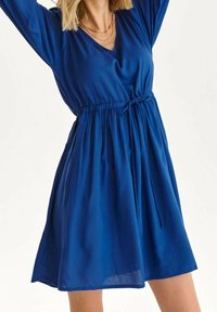 Sininen mekko, joka on valmistettu kevyestä kankaasta, jossa on V-pääntie, pitkät hihat, nyöritetty vyötärö ja A-linjainen siluetti.