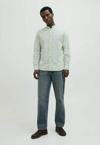 Homem em pé, vestindo uma camisa de botão verde claro, jeans azuis largos e sapatos castanhos escuros, contra um fundo liso claro.