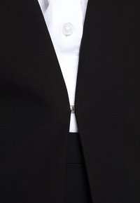 Blazer noir fermé par une boucle argentée sur une chemise blanche avec un bouton visible au col.