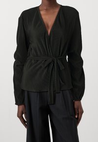 Blouse plissée vert foncé avec un décolleté en V profond, manches longues et un détail de ceinture à nouer à la taille. Texture lisse, tissu fluide.