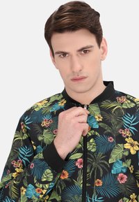 Jachetă bomber florală cu fundal negru, având frunze și flori vibrante în nuanțe de verde, galben, roz și albastru. Închidere cu fermoar.
