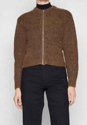 Personne portant un pull brun duveteux à fermeture éclair sur un haut noir, assorti d’un pantalon noir taille haute, debout devant un fond uni.
