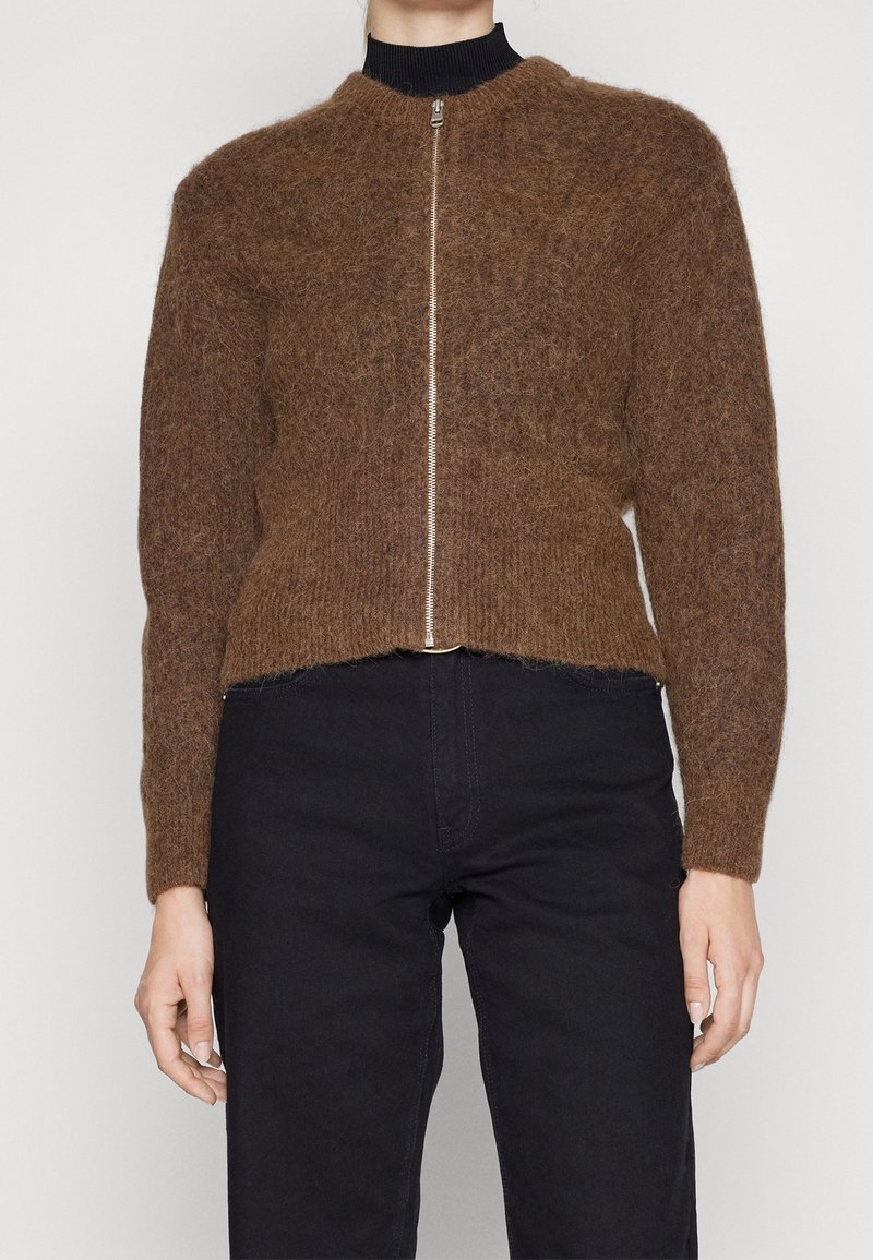 Personne portant un pull brun duveteux à fermeture éclair sur un haut noir, assorti d’un pantalon noir taille haute, debout devant un fond uni.