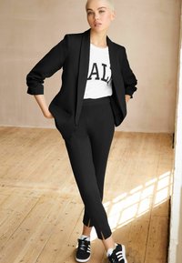 Svart skräddarsydd blazer, vit t-shirt med iögonfallande "AL"-tryck, svarta åtsittande byxor och svarta sneakers med vita ränder. Bakgrund av trägolv.
