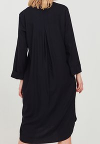 Robe noire de longueur genou en tissu doux, dotée d'une coupe décontractée, de manches trois-quarts et d'un pli dans le dos pour un détail supplémentaire.