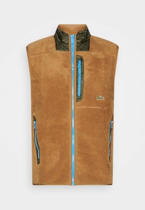 Veste en polaire marron avec des fermetures éclair bleues, un liseré vert sur le col et les poches, avec un petit logo crocodile sur la poitrine.