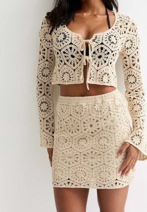 Vrouw draagt een beige gehaakt tweedelig outfit met een crop top met lange mouwen en strik aan de voorkant en een hoge taille minirok, waar zwarte onderkleding onder zichtbaar is.