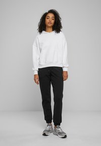 Weißer Hoodie mit einer Kängurutasche, kombiniert mit schwarzen Jogginghosen. Das Outfit hat einen lässigen Schnitt und strukturierter Stoff. Graue Sneaker runden den Look ab.