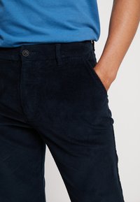 Tummansininen korvapuistojobo, jossa on teksturoitu viimeistely, sivutaskut ja yksittäinen napitus vyötäröllä.