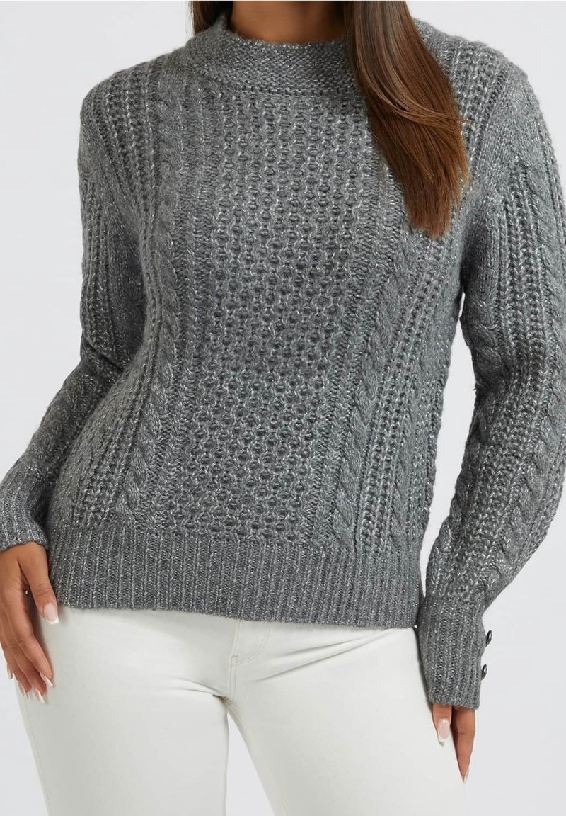 Femme portant un pull en maille torsadée gris avec des poignets boutonnés et un pantalon blanc, vue du cou jusqu'aux hanches.