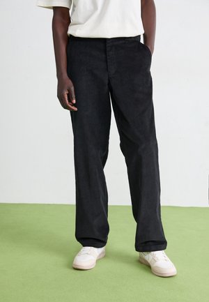 Person wearing black wide-leg pants, white sneakers, and a white short-sleeve shirt, standing on green floor against a plain light wall.