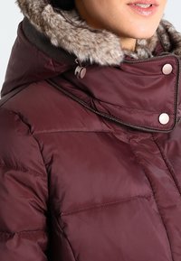 Burgunderfarvet pufjakke med hætte foret med faux pels, trykknapper og kviltet struktur. Har justerbar snøre til en tætsiddende pasform.