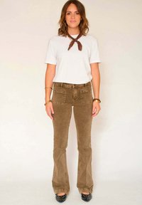 Pantalon évasé en velours côtelé marron associé à un t-shirt blanc et un foulard marron autour du cou, avec des poches et un tissu texturé. Chaussures à talons noires.