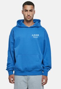 Modra jopa s kapuco, velikim sprednjim žepom in belim napisom. Iz mehke tkanine s sproščenim krojem, opremljena z rebrastimi manšetami in spodnjim robom.