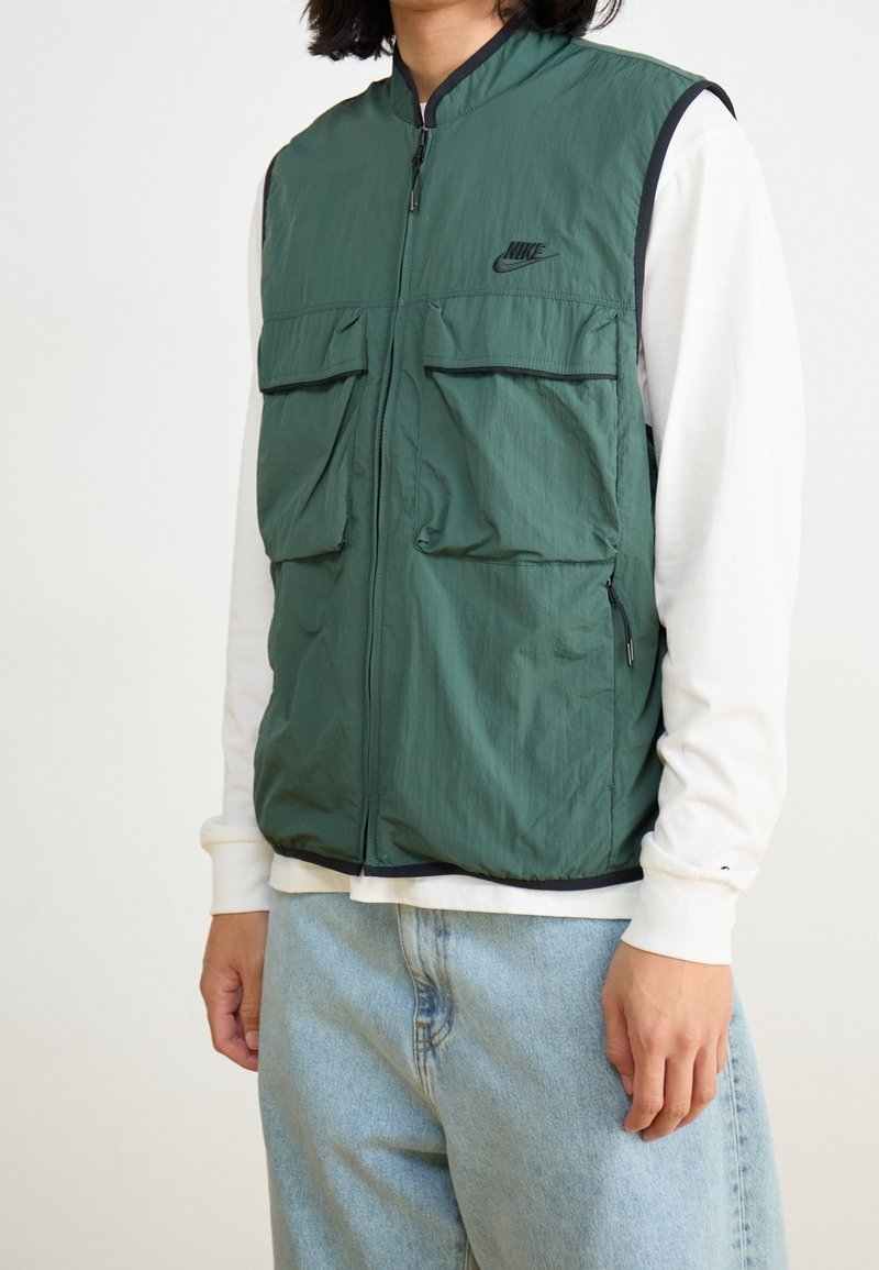 Zelená lehká vesta se zipem, dvěma velkými předními kapsami a malým logem. Nositelná přes bílé tričko s dlouhým rukávem, minimalistický design.