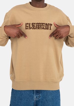 Person trägt beiges Sweatshirt mit dem Wort "ELEMENT" in braunem Animal-Print-Schriftzug und zeigt auf den Text im Brustbereich.