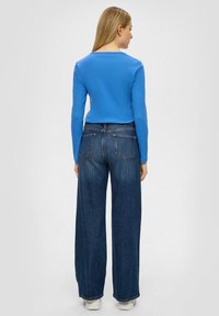 Blauwe longsleeve top met een ronde hals, gecombineerd met wijd uitlopende donkere denim jeans met achterzakken en een rafelige zoom. Witte sneakers.