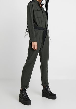 Personne portant une combinaison longue verte foncée avec poches poitrine, ceinture noire, manches retroussées et bottines à plateforme noires lacées.