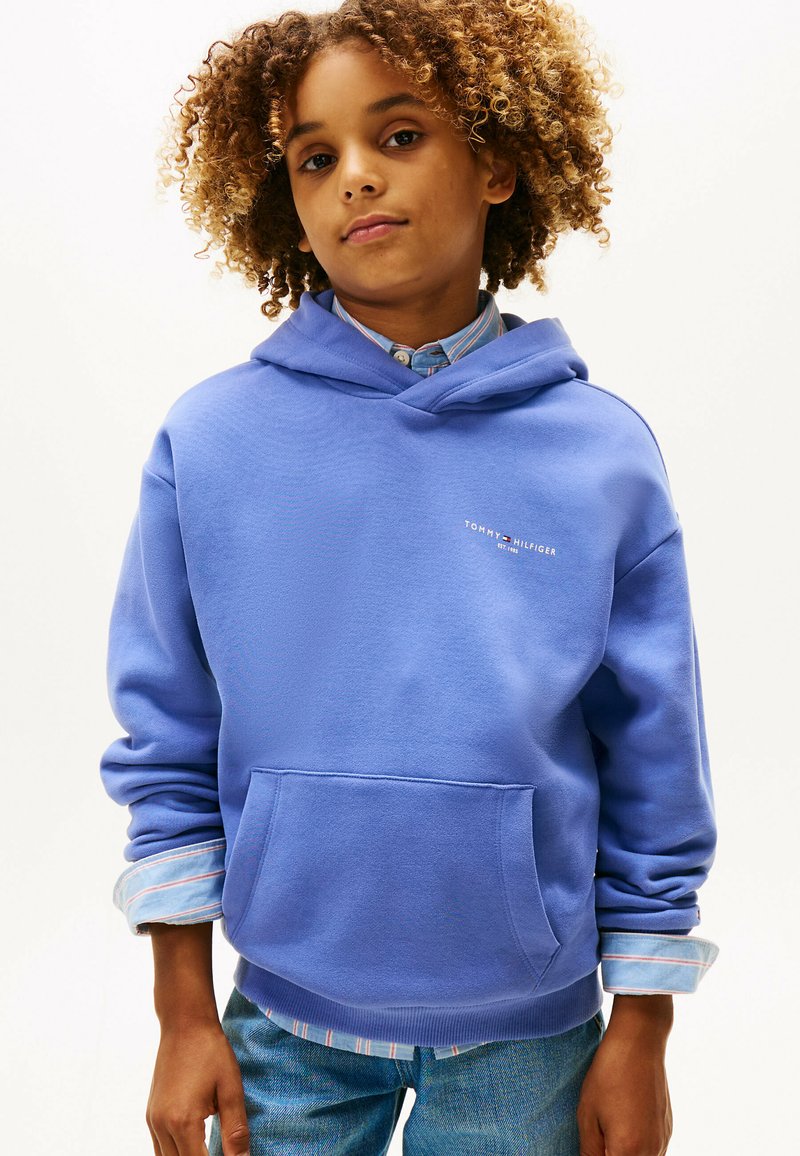 Kind met krullend haar, gekleed in een blauwe Tommy Hilfiger hoodie over een gestreept shirt en een spijkerbroek, staand tegen een effen achtergrond.