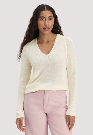 Woman with long curly hair wearing a cream V-neck sweater and light pink high-waisted pants, standing against a plain background.