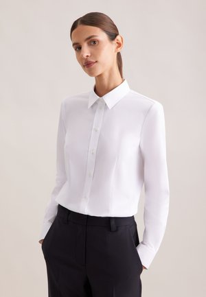 Frau mit braunen Haaren in einem weißen Hemd mit Knöpfen und schwarzen Hosen, die mit den Händen in den Taschen vor einem schlichten beigen Hintergrund steht.