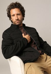 Zwarte ritsjack met gequiltte panelen, hoge kraag en lange mouwen, gedragen over een donkerbruin top, gecombineerd met beige broeken.