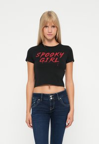 Joven mujer vestida con una camiseta corta negra con el texto rojo "SPOOKY GIRL" y unos vaqueros azul oscuro, de pie contra un fondo blanco liso.