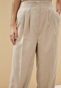 Pantalones plisados beige con un corte ajustado, que cuentan con una cintura alta y bolsillos laterales, hechos de una tela suave y ligera.