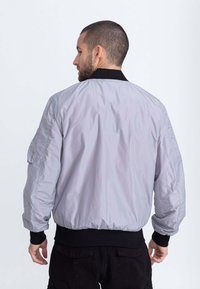 Giacca bomber grigia chiara con colletto e polsini a coste neri. Presenta una tasca sulla manica e una cintura elastica. Materiale liscio e leggero.