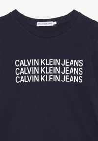 Laivastonsininen puuvillapaita, jossa on toistuva valkoinen "CALVIN KLEIN JEANS" -teksti. Pyöreä kaula-aukko ja sileä pinta. Tagi näkyvissä kaula-aukossa.