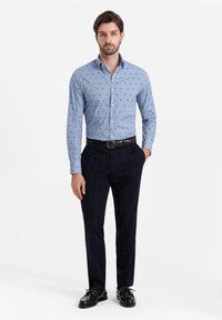 Camisa de botón azul claro con patrones oscuros dispersos, combinada con pantalones a rayas oscuros y zapatos negros. Cuello simple y mangas largas.