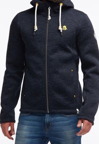 Ciemnoniebieska bluza zapinana na zamek, wykonana z teksturowanej tkaniny, z białymi sznurkami, żółtymi akcentami i bocznymi kieszeniami. Luźny krój z prążkowanymi mankietami.