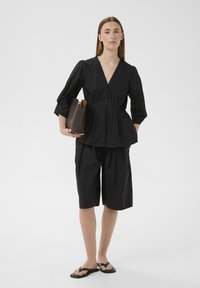 Femme portant un chemisier noir à col en V boutonné avec des manches bouffantes, un short noir au genou, tenant une pochette marron texturée, et des sandales noires.