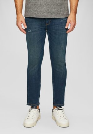 Jeans en denim bleu marine avec une coupe slim, légèrement déchirés, et des poignets retroussés. Associés à des baskets blanches avec des accents noirs.