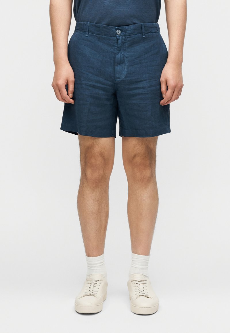 Σορτς από λινό σκούρου μπλε με κούμπωμα με κουμπί, επίπεδη μπροστινή όψη και πλαϊνές τσέπες. Συνδυασμένα με ανοιχτόχρωμα αθλητικά παπούτσια και κάλτσες.