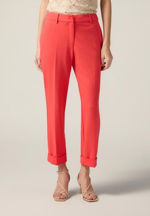 Koraal kleurige pants met een getailleerde pasvorm, voorzien van een knoopsluiting, opgerolde boorden en een gladde stoftextuur. Draagt met naaktkleurige hoge hakken.