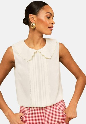 Femme portant un haut blanc sans manches à col festonné et un pantalon à carreaux rouges et blancs, mains dans les poches, tête tournée sur le côté.