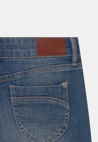 Denim-Jeans mit einem mittelblauen Waschung. Mit einem braunen Lederpatch, auf dem "Pepe Jeans London" steht, und sichtbaren Nähten, sowie einem Detail mit seitlichen Taschen.