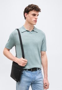 Νεαρός άνδρας που φοράει ανοιχτοπράσινο πόλο με κοντά μανίκια και ανοιχτό μπλε τζιν, κρατώντας μια μαύρη τσάντα χιαστί στον αριστερό του ώμο.