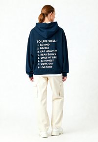 Hanorac bleumarin cu text alb care enumeră opt principii de bunăstare. Croială lejeră cu o glugă mare, asortat cu pantaloni cargo crem. Textură de material neted.