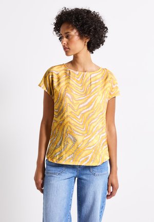 Maglietta a maniche corte gialla con un motivo ondulato in arancione e grigio. Realizzata in tessuto leggero con una vestibilità ampia. Abbinata a jeans azzurri chiari.