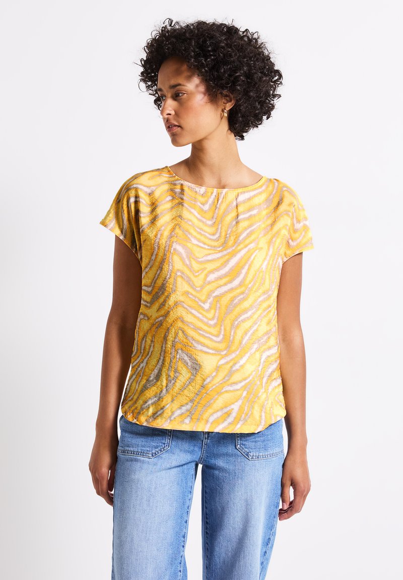 Top amarillo de manga corta con un patrón ondulado en naranja y gris. Hecho de una tela ligera con un ajuste holgado. Combinado con jeans de color azul claro.