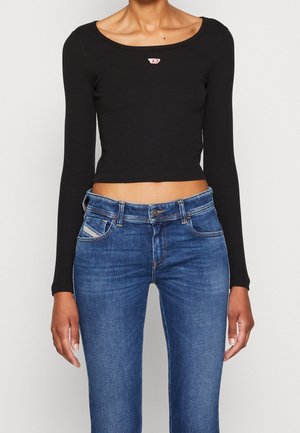 Femme portant un crop top noir moulant à manches longues avec un petit logo rouge et blanc sur la poitrine et un jean bleu taille haute.